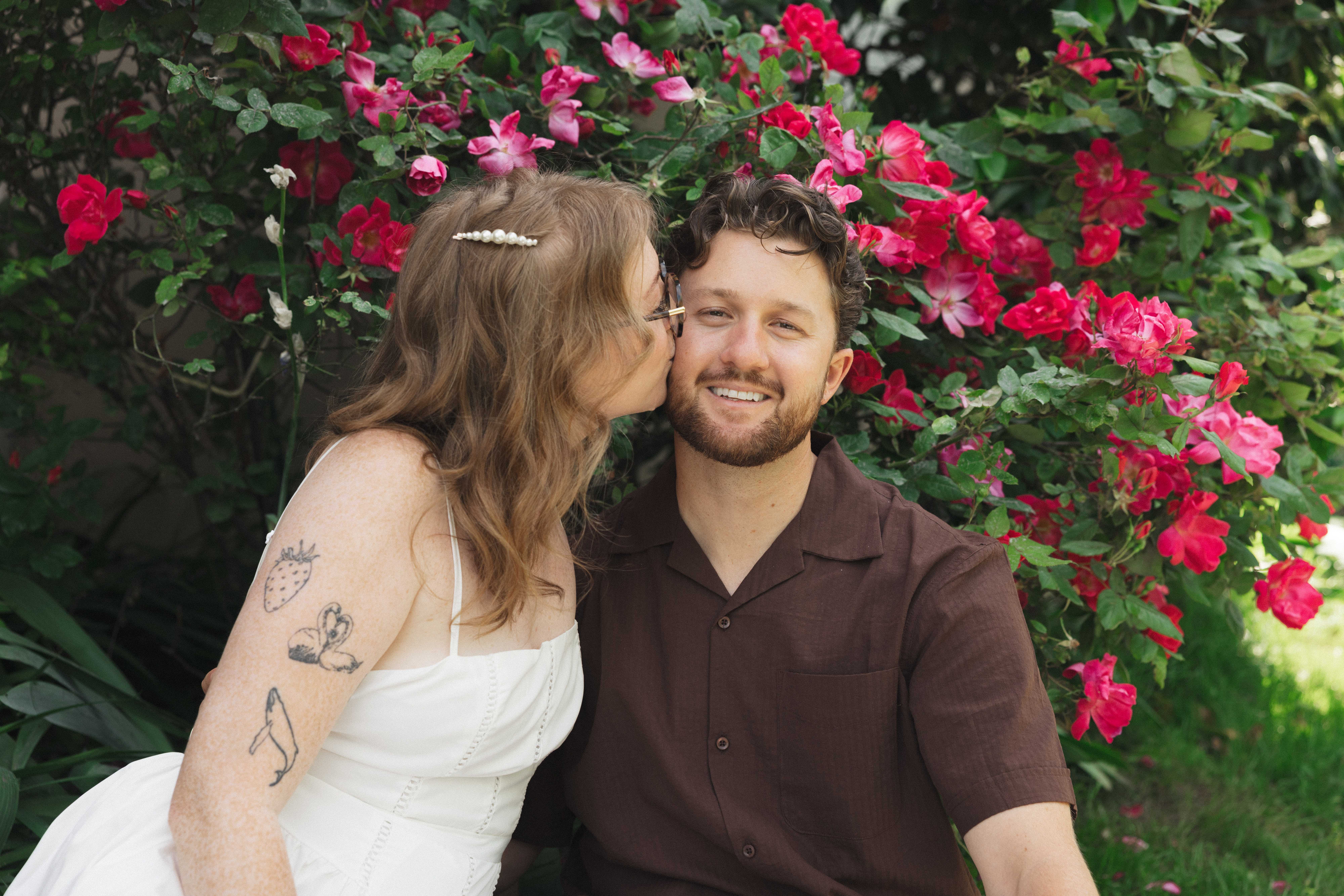 Madison kissing Evan on the cheek in front of a blooming rose bush